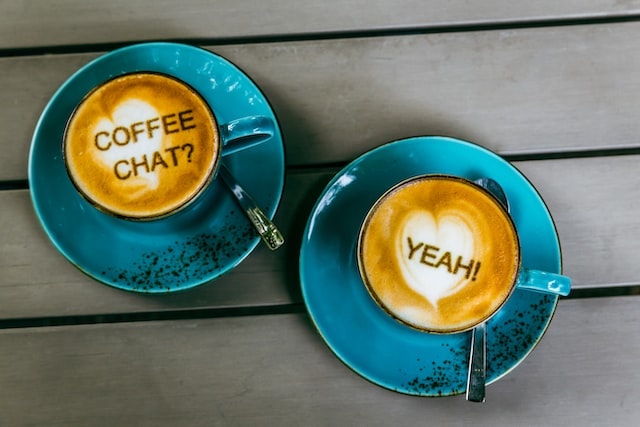 Two cups of freshly brewed coffee (1 cappuccino, 1 wet cappuccino) on a table, with a text on the coffee foam: "Coffee Chat? YEAH!". The print was applied to the milk foam by a professional barista coffee printer. Photo by Frank Leuderalbert.